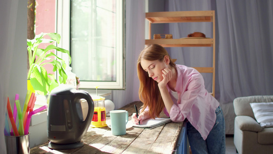 年轻妇女站在厨房的木桌旁喝茶和做笔记视频