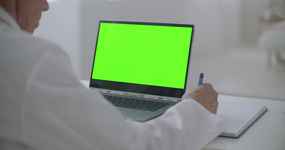医生正在看笔记本电脑在诊所办公室桌上用绿色屏幕检查视频