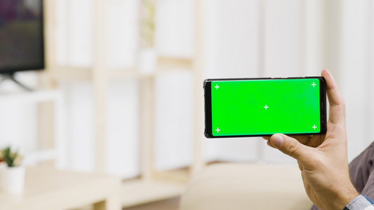 在现代而明亮的客厅里男人手里握着一个绿色的屏幕电话视频