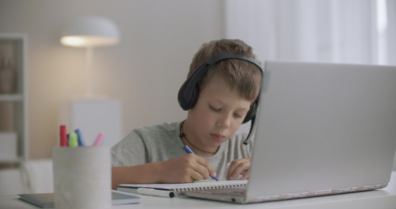 手头上戴耳机和桌上手提笔记本电脑的男孩在孤立时期在视频