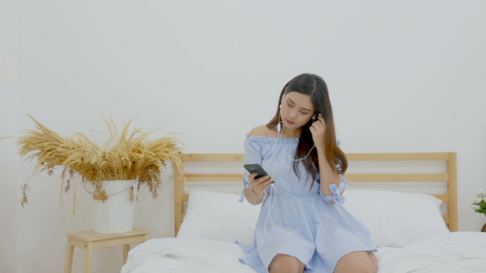 美丽的女人坐在床上用手机应用程序中的耳机听音乐视频