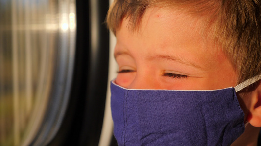 男孩在乘火车时望着窗外。戴着棉质面罩的孩子的特写镜头。阳光照在乘客脸上。运输中戴防护面罩的儿童。小旅行者的面部视频