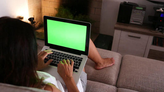 一名年轻妇女在她的笔记本电脑上工作,在家用绿色屏幕做画,坐在她客厅的沙发上视频