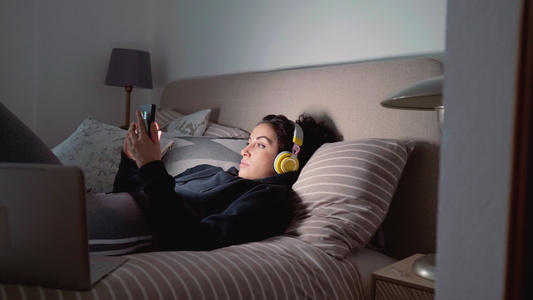 戴着耳机躺在床上使用智能手机的年轻女性视频