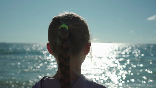 金发小女孩的特写，扎着辫子，她看着日落时闪闪发光的海水。风吹过她的头发。她在做梦视频