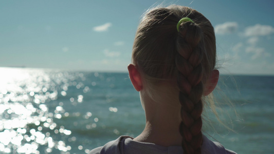 用辫子看着在日落时闪闪发光的海水的金发小女孩的特写。风吹过她的头发。梦想着未来视频