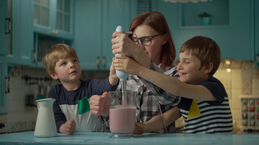 幸福的母亲和两个孩子在家里的蓝色厨房里用搅拌机烹制草莓、香蕉、纯素牛奶和酸奶的冰沙。男孩倒出牛奶，开始搅拌鸡尾视频