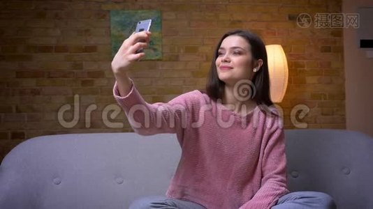 一位年轻漂亮的黑发白种人女性在电话上自拍，坐在沙发上微笑的特写镜头。视频