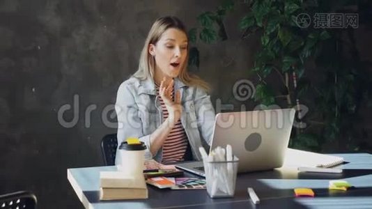 有魅力的年轻女性正在电脑上通过Skype聊天，而坐在现代办公室的桌子上。 她在说话视频