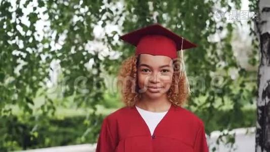 穿着红色长袍和灰泥板的漂亮非裔美国女毕业生站在户外微笑着的肖像视频