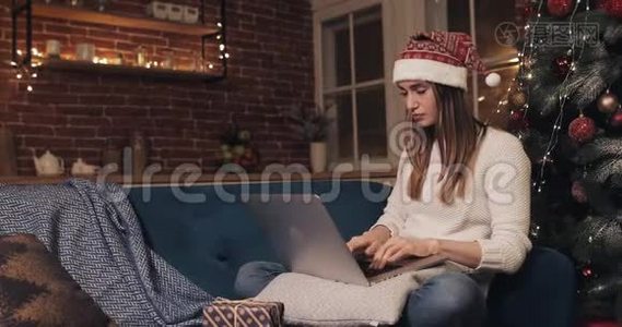 迷人的白种人女孩坐在圣诞树附近使用她的笔记本电脑思考和命令现在。 礼品盒出现在视频