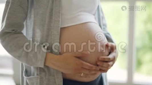 期待的母亲正在抚摸她的大婴儿肿块，站在室内，背景是大窗户。 快乐的怀孕，人们视频