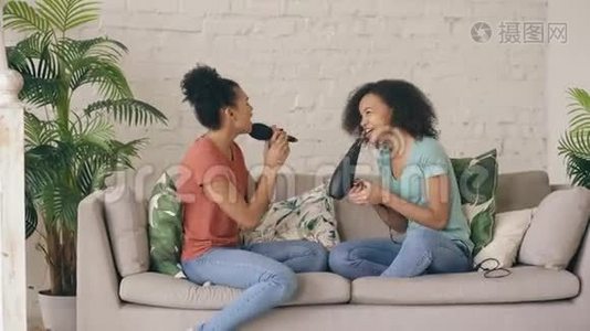 混合种族年轻有趣的女孩跳舞唱歌，吹风机和梳子坐在沙发上。 姐妹享受休闲生活视频