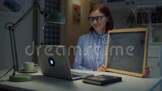 年轻微笑的30多岁的女人戴着眼镜，拿着干净的空粉笔板，看着家里的笔记本电脑。 在线教育程序视频