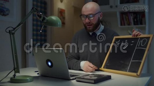 长着胡子的30多岁的男老师戴着眼镜在粉笔板上写数学，用白色粉笔看着家里的笔记本电脑。 网上在线视频