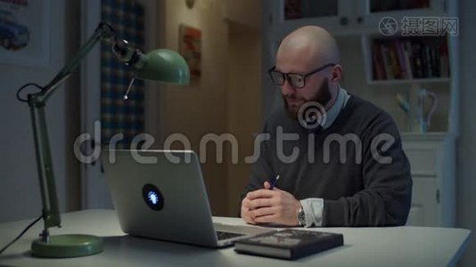 30多岁的大胡子戴眼镜的男人在家里打视频电话时，在笔记本电脑上情绪化地交谈。 在线教育过程。 侧视视频
