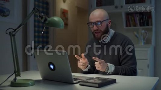 30多岁的大胡子戴眼镜的男人在家里打视频电话时，在笔记本电脑上情绪化地交谈。 在线教育过程。 侧视视频