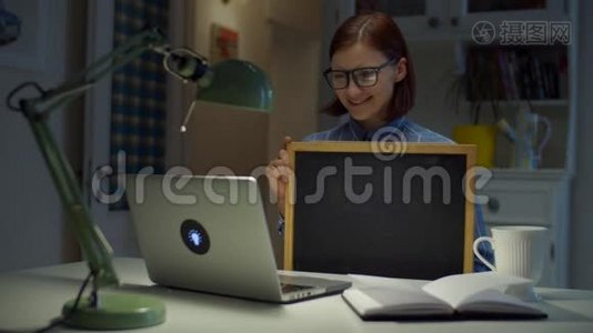 30多岁的年轻女性戴着眼镜，手里拿着干净的空粉笔板，看着家里的笔记本电脑。 在线教育过程。 工作视频