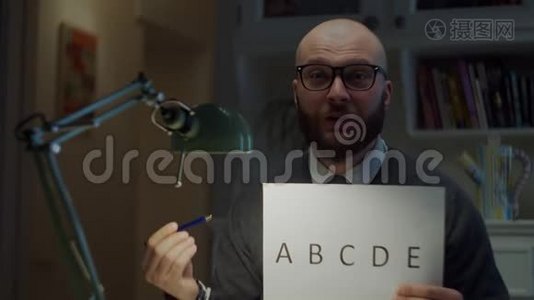 30多岁的留着胡子的男人戴着眼镜，在家里的摄像机前展示一张纸和字母表。在线教育过程视频