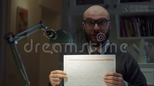 一个30多岁的留着胡子的戴眼镜的男人在家里用相机展示一张有图表的纸。在线教育过程。网络摄像机视图视频