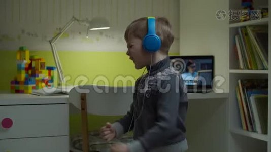 戴着彩色耳机的学龄前男孩在家里跳舞，背着笔记本电脑和女老师上在线课。电子学习视频