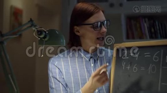 30多岁的女人戴着眼镜在粉笔板上写数学，用白色粉笔在家里看着相机。 在线教育程序视频
