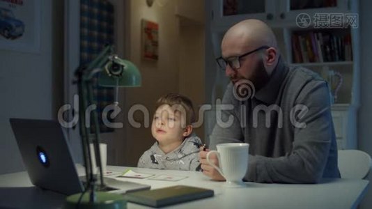 年轻的30岁的父亲和学龄前儿童在家做在线家庭作业。 大胡子男和小男孩在网上学习视频