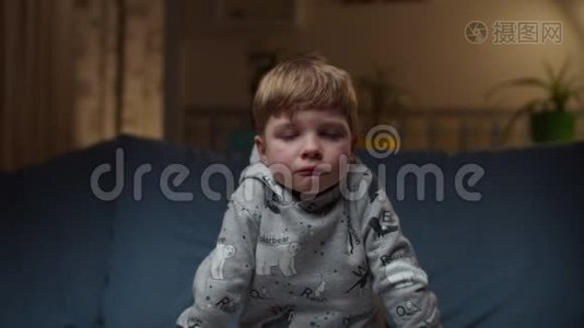 小男孩坐在家里的沙发上的肖像。 悲伤的孩子`脸缓慢地看着摄像机。视频