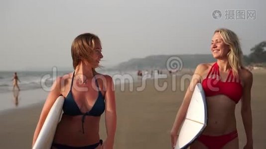 两个穿着泳衣的年轻女人带着冲浪板在海滩上散步。 慢动作视频