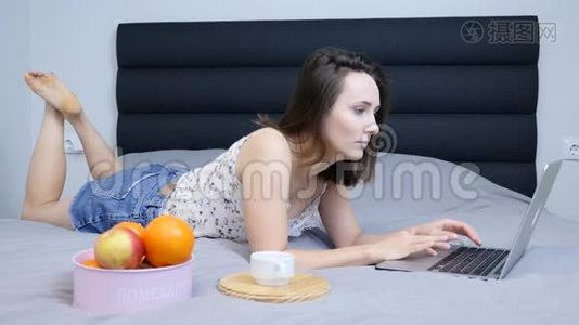 女千禧看着笔记本电脑屏幕躺在客厅的床上。 穿短裤的年轻女子在笔记本电脑上上网聊天视频