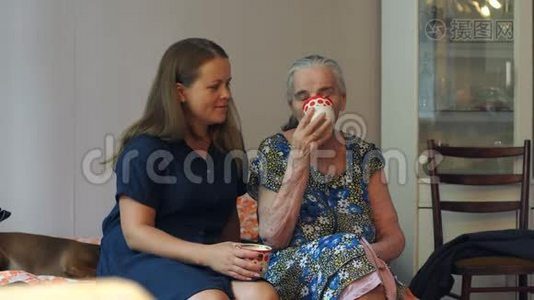 年轻的女人和她八十岁的祖母在家里的沙发上喝茶。视频