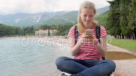 一个女孩和她的智能手机坐在海边的山脉和树木。视频