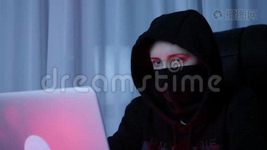 在电脑上关闭戴黑色面罩和引擎盖的女性面部密码。 黑客的脸看着笔记本电脑屏幕和编码网络视频