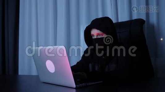 女黑客坐在黑暗的房间里，在笔记本电脑键盘上打字。 戴着黑色面具和罩的女人在屏幕上打电话视频
