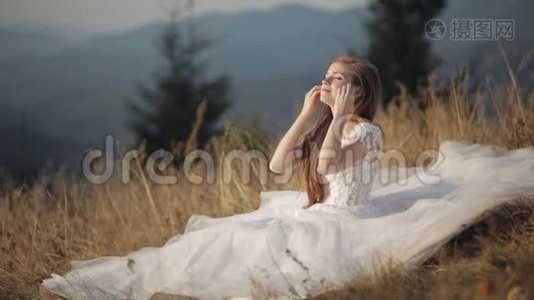 美丽可爱的新娘穿着婚纱坐在山坡上的草地上视频