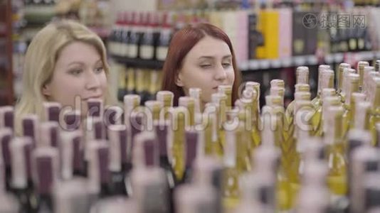 年轻的红发女人，带着绿色的眼睛看着货架上的酒，像一个开朗的金发朋友加入她的行列。 快乐的成人视频