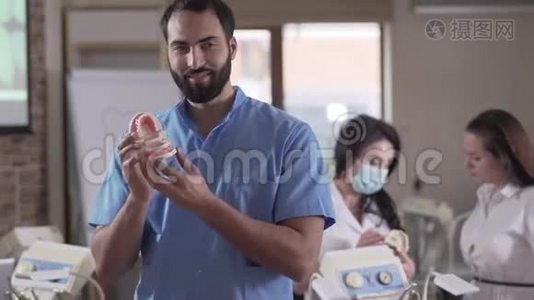 正面的中东男牙科医生在镜头前微笑的肖像，手里拿着牙齿，同事在镜头前视频