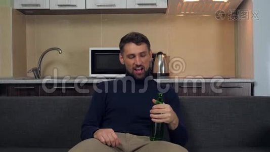 一个穿着深蓝色套衫和灰色裤子的年轻人坐在沙发上，喝着啤酒，微笑着看电视上的娱乐节目视频