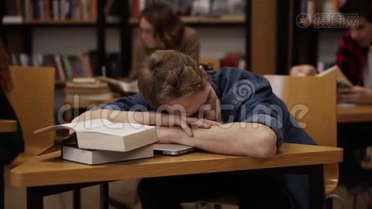 年轻的欧洲学生在大学图书馆或班级睡在桌子上，上面有一堆书。 同学和书架视频