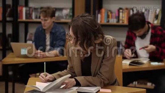 一个穿着棕色夹克的古丽的欧洲女孩坐在大学图书馆里写论文，准备考试，然后看上去视频