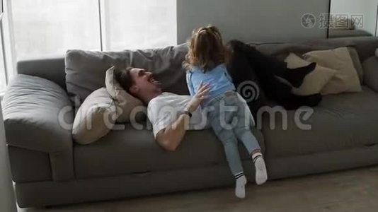 快乐的家庭爸爸和小女儿一起玩，躺在沙发上，爸爸挠着女孩的痒，在他的怀里长大，还有视频