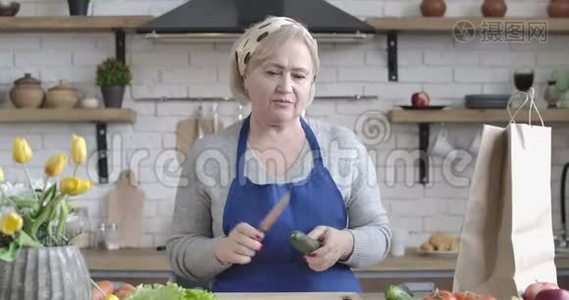 自信的高加索族女人在厨房里用黄瓜和刀子说话的肖像。 中年退休人员视频