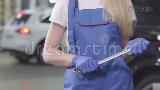 无法辨认的金发女人穿着蓝色工作袍和手套拿着扳手。 年轻的女汽车修理工站在视频