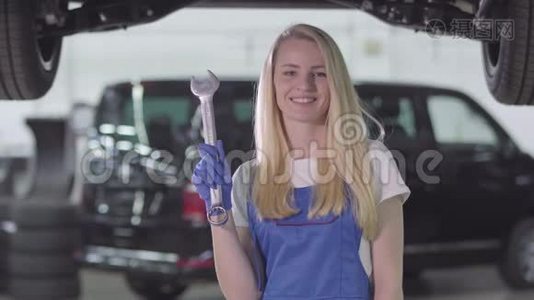 微笑的女汽车机械师，穿着工作服，拿着扳手，伸出拇指。 金发白种人女人看着摄像机视频