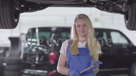 年轻的女汽车修理工穿着工衣站在修理厂的车下，看着摄像机，微笑着的画像视频