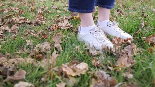 女人`脚穿着白色的运动鞋在绿草上，落叶。 静态特写镜头，秋季自然视频