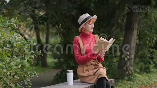 戴着帽子、穿着毛衣的年轻红发女郎在长椅上看书，喝着热咖啡视频