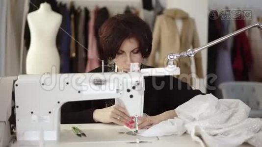 一位成年女裁缝的肖像在一台缝纫机上工作，工作在一件白色手工制作的连衣裙上。 时尚服装和时装视频