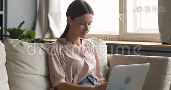 年轻女士用笔记本电脑打字坐在家里的沙发上视频