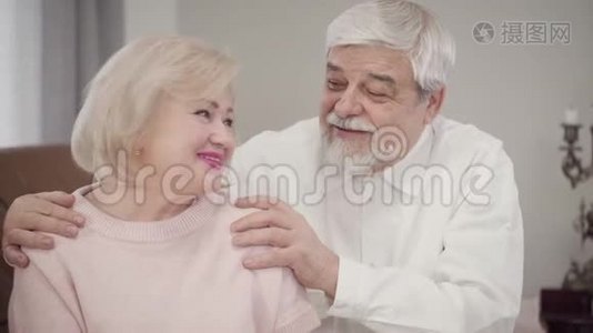 年长的高加索夫妇互相交谈，看着相机，微笑。 已婚退休人员在室内摆姿势视频